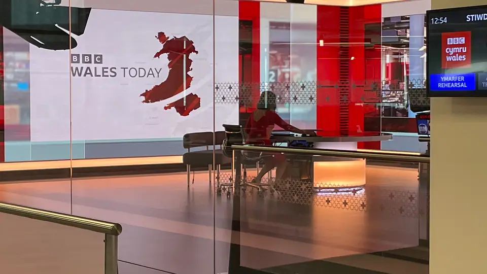 Estúdio de telejornal com bancada e telas ao fundo